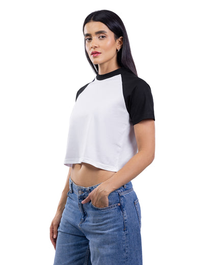 A woman modeling a short-sleeve crop top with a white body and black raglan sleeves, paired with blue jeans, against a plain white background.