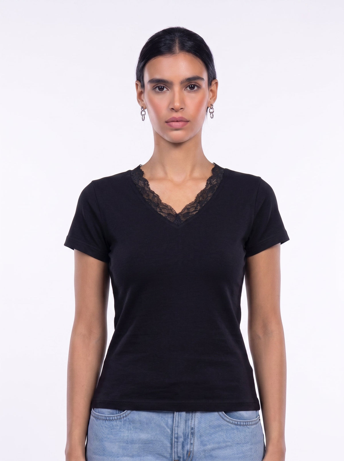 A woman modeling a black short-sleeve V-neck t-shirt with a black lace trim along the neckline, paired with light blue jeans, against a plain white studio background.