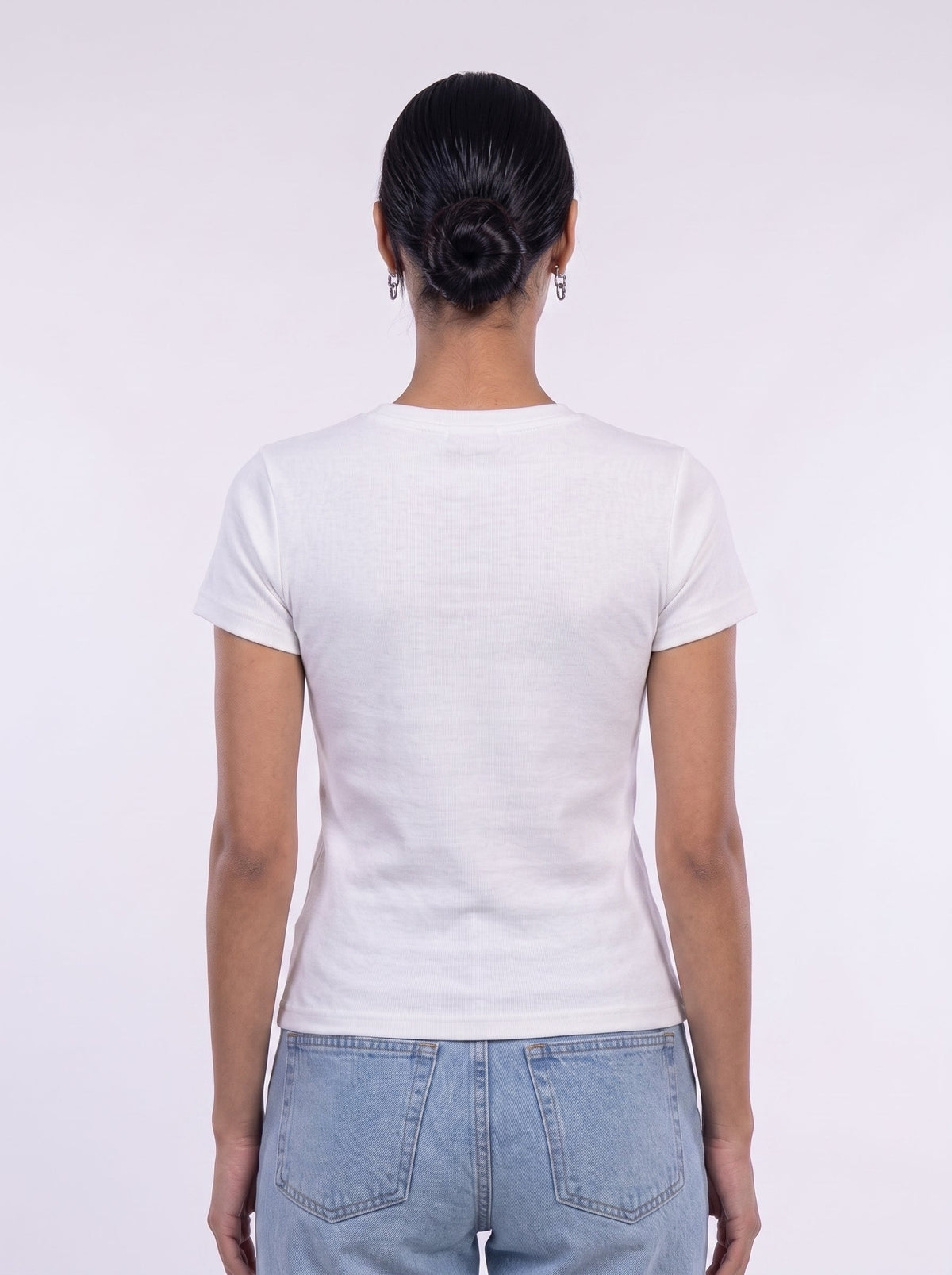 A woman viewed from behind wearing a plain white short-sleeve crewneck t-shirt and light blue jeans against a light gray studio background.