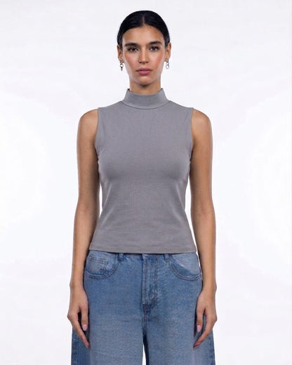 A woman modeling a sleeveless gray mock neck top paired with blue jeans against a plain white studio background.