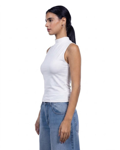 A woman in profile wearing a white sleeveless mock neck top and light blue jeans, photographed against a solid white background.