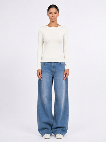 A model stands against a plain white studio background wearing a fitted off-white long-sleeve top, wide-leg light blue jeans, and white shoes.