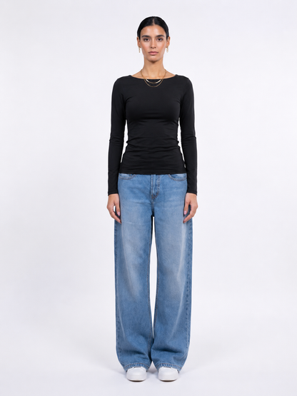 A model stands against a plain white studio background wearing a fitted black long-sleeve top, wide-leg light blue jeans, and white sneakers.